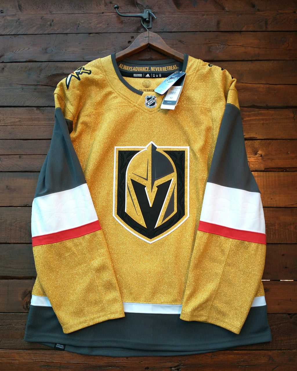 A Vegas Golden Knights hockey jersey in gold, black, and white, hanging on a wooden hanger against a dark wood plank wall. The jersey features the team's helmet logo on the front.