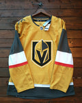 A Vegas Golden Knights hockey jersey in gold, black, and white, hanging on a wooden hanger against a dark wood plank wall. The jersey features the team's helmet logo on the front.