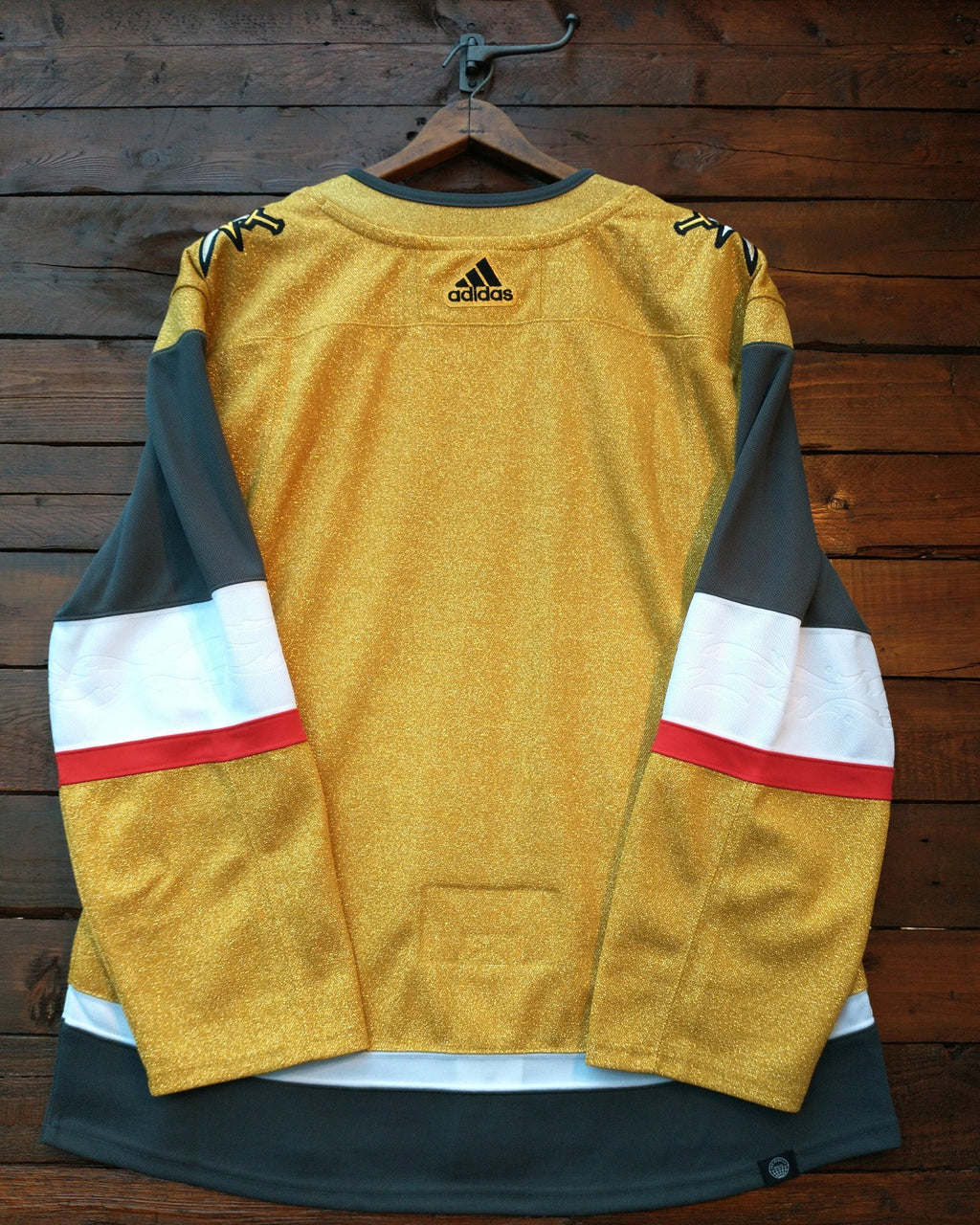 A gold Adidas hockey jersey with dark gray and white sleeve stripes and red accents, hanging on a wooden hanger against a dark wood plank wall.
