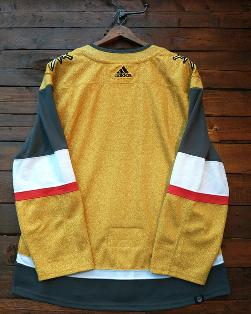 A gold Adidas hockey jersey with dark gray and white sleeve stripes and red accents, hanging on a wooden hanger against a dark wood plank wall.
