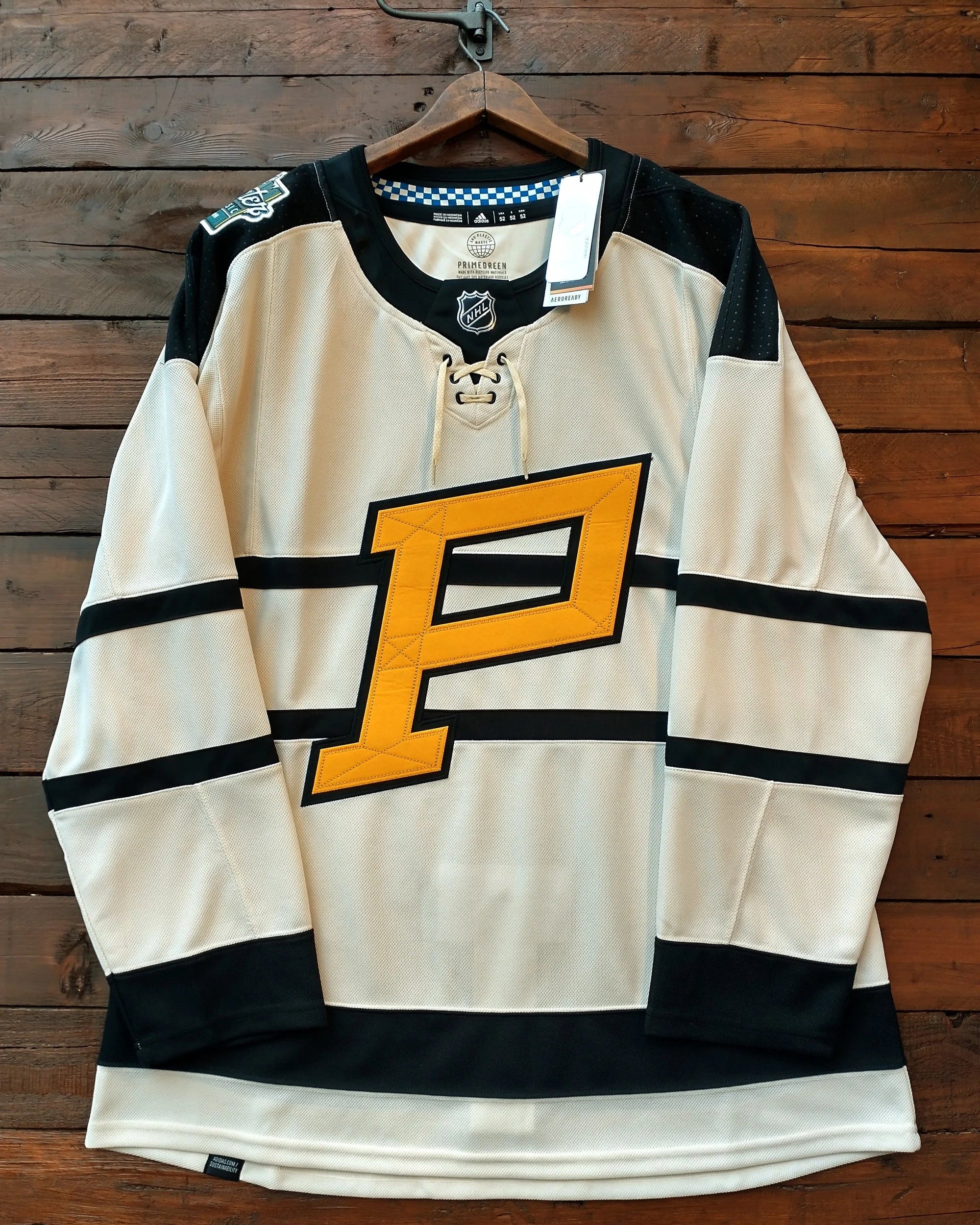 A Pittsburgh Penguins hockey jersey hanging on a wooden hanger against a dark wood plank wall. The jersey is cream with black shoulder, sleeve, and hem stripes, and features a large yellow and black 'P' logo on the chest.