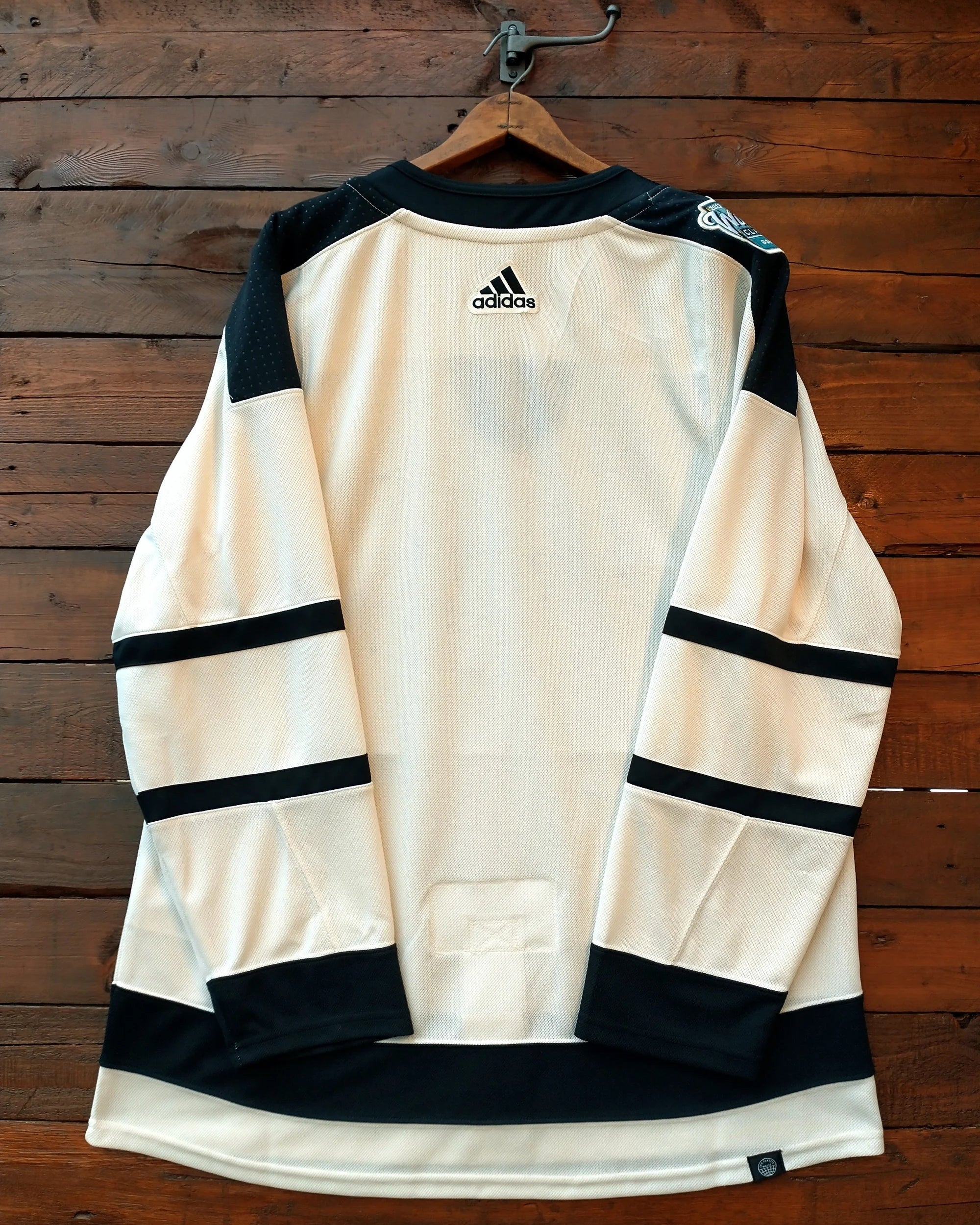 An Adidas hockey jersey in cream and black hanging on a wooden hanger against a dark wood plank wall. The Adidas logo is visible on the upper back.