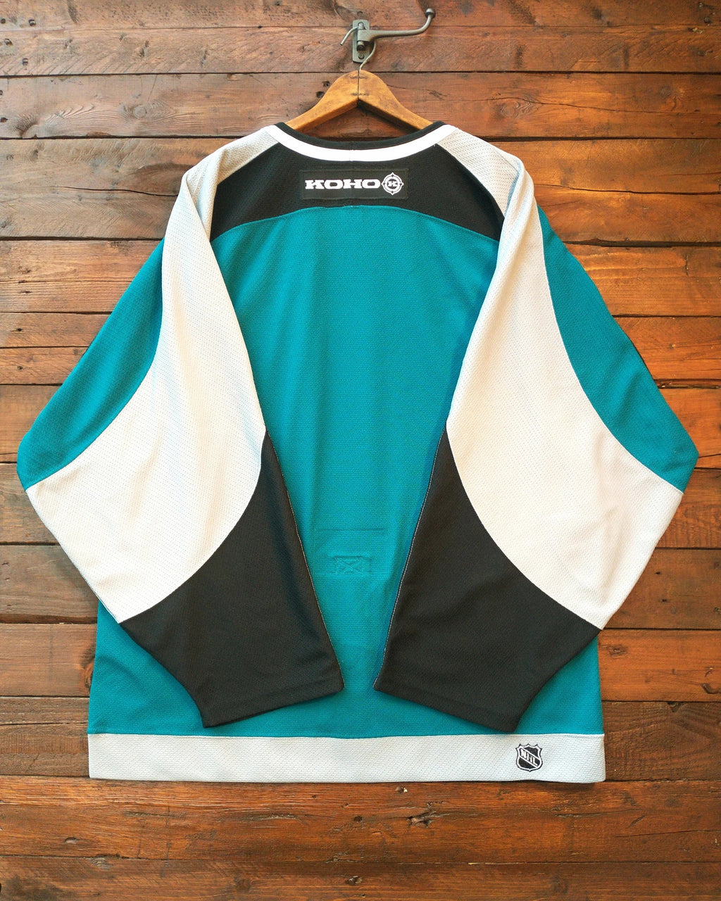 A Koko hockey jersey in teal, white, and black, shown hanging on a wooden hanger against a rustic wooden plank wall. The back of the jersey features the Koko logo in white on a black panel at the upper back.
