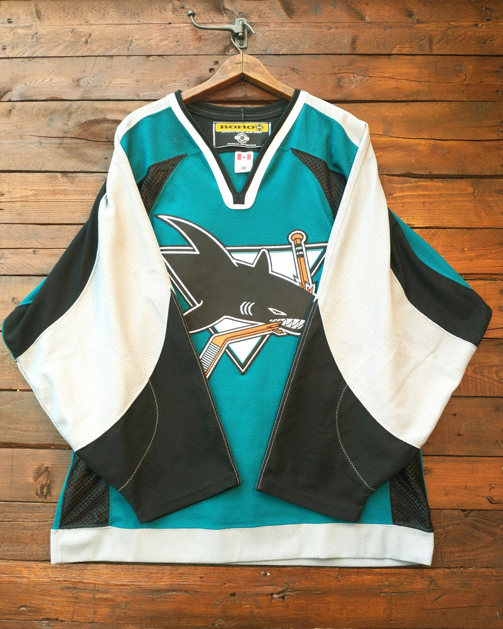 A San Jose Sharks hockey jersey in teal, black, and white, hanging on a wooden hanger against a rustic wooden plank wall. The jersey features the team's shark logo on the front.