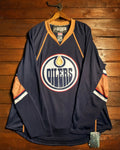 An Edmonton Oilers long-sleeve hockey jersey hanging on a wooden hanger against a rustic wooden plank wall. The jersey is navy blue with orange and white accents, featuring the team's circular logo on the chest.