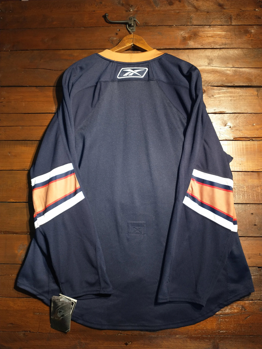 A navy blue Reebok hockey jersey hanging on a wooden hanger against a rustic wooden plank wall. The jersey features white and orange stripes on the sleeves and a white Reebok logo patch on the back neck.