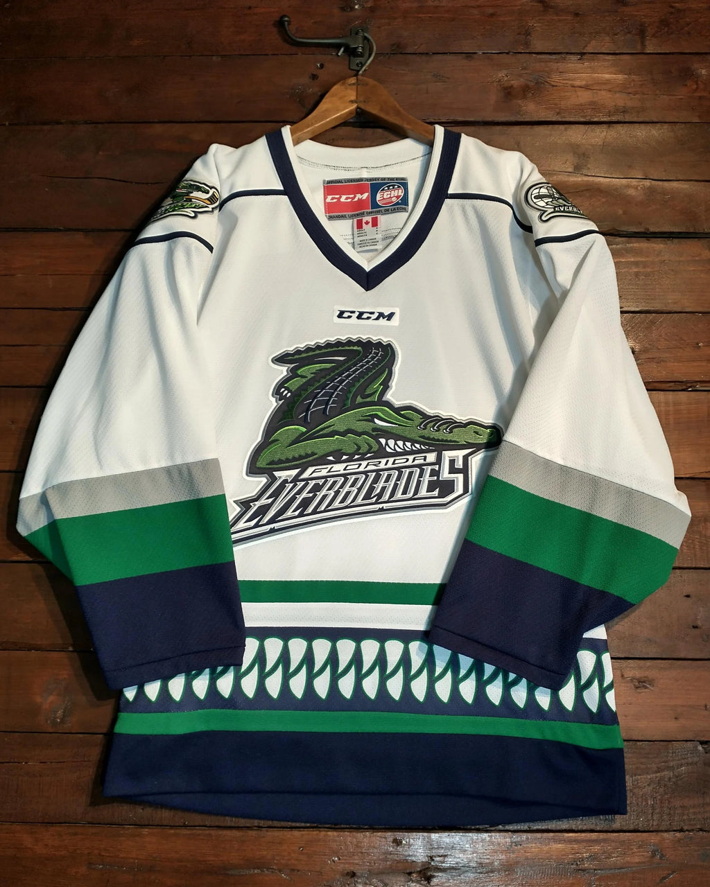 A white CCM Lake Superior Northwoods AHL hockey jersey with green and navy blue accents, hanging on a wooden hanger against a dark wood plank wall.