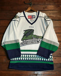 A white CCM Lake Superior Northwoods AHL hockey jersey with green and navy blue accents, hanging on a wooden hanger against a dark wood plank wall.