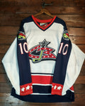 A vintage hockey jersey with a white, navy, and red color scheme, featuring the number 10 and a star-and-stripe logo on the front, hanging on a wooden hanger against a rustic wood plank wall.