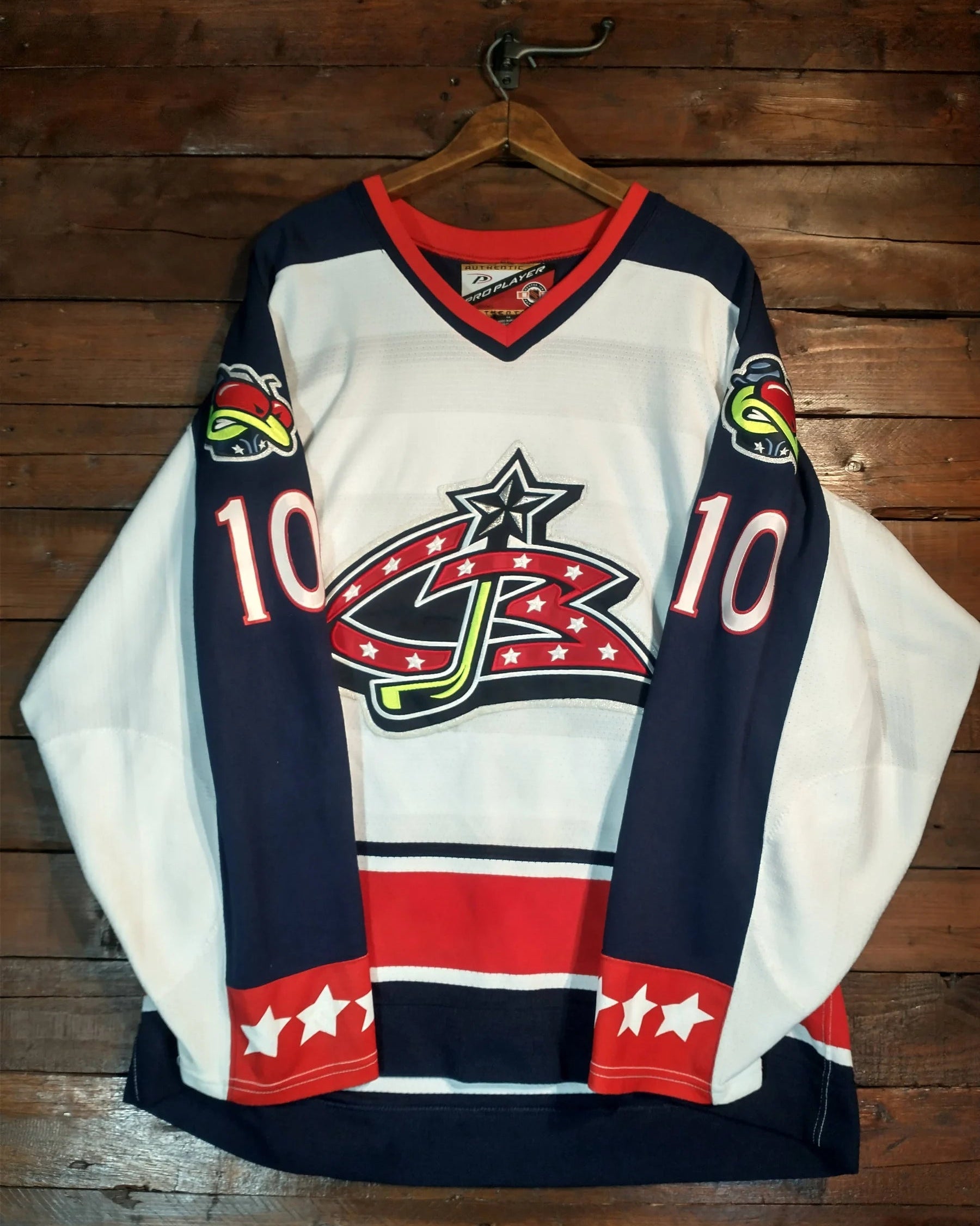 A vintage hockey jersey with a white, navy, and red color scheme, featuring the number 10 and a star-and-stripe logo on the front, hanging on a wooden hanger against a rustic wood plank wall.